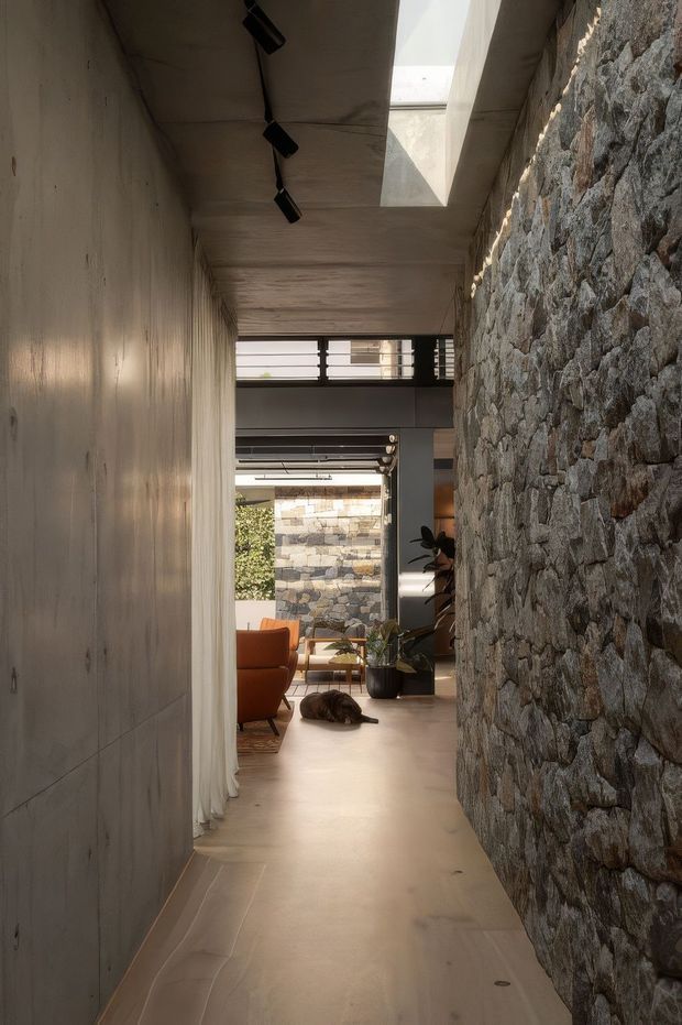 A narrow contemporary interior corridor leading into a living area. The passage has a raw concrete ceiling and wall on one side, a prominent rough-faced stone feature wall on the other, recessed linear track lighting, and a rectangular skylight. A light timber-toned floor leads toward an open lounge with orange upholstered seating, plants, and a visible stone courtyard wall beyond.