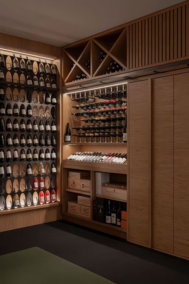 A dark, refined wine storage and display area with extensive custom timber cabinetry. Shelves on the left hold many wine bottles, while the central illuminated display includes horizontal bottle racks, wine glasses, boxed stock, and additional bottles. Upper open cubbies feature geometric cross-bracing, and the right side has tall slatted and paneled timber doors. Warm integrated lighting creates an intimate, premium atmosphere.