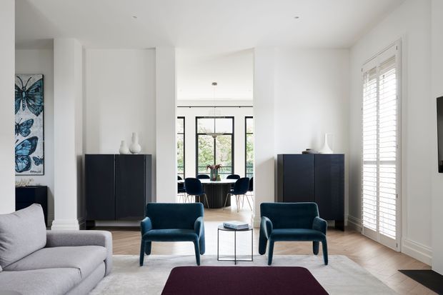 Bright, refined contemporary living room with white walls, pale timber flooring, a large light rug, deep blue upholstered armchairs, and a partially visible sofa. Black built-in or freestanding storage cabinets line the walls, while full-height white plantation shutters frame the windows. The open-plan dining area beyond includes a dark dining table and blue chairs, with large glazed doors bringing in natural light. Abstract butterfly artwork and restrained decorative objects add subtle contrast.