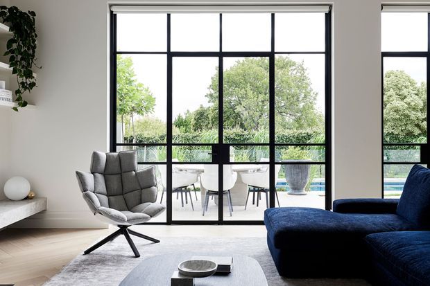 A bright contemporary living room with large black-framed floor-to-ceiling grid glazing opening to an outdoor terrace and garden. The room features a deep navy-blue sectional sofa, a sculptural grey lounge chair, a low light-grey coffee table, pale timber flooring, a neutral rug, and a minimalist white built-in ledge with a potted plant. The glazing and restrained palette create a refined indoor-outdoor connection.