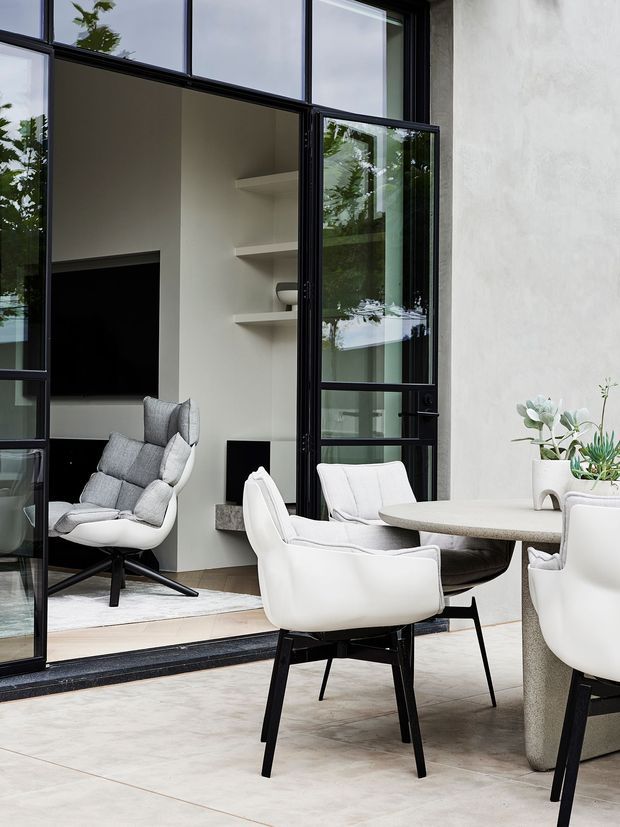 A refined contemporary dining and living area with pale stone-look floor tiles, a rounded light dining table, and sculptural white upholstered chairs with dark timber or black legs. Large black-framed glazed doors and windows open toward a sunken living room containing a television, built-in shelving, and a pale lounge chair. Neutral plaster walls, soft furnishings, and greenery create a calm, minimalist atmosphere.