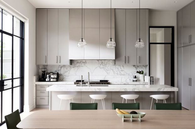 A refined contemporary kitchen and dining area with full-height grey cabinetry, a large marble-look splashback, a wide island with seating for four, and three glass pendant lights. Black-framed glazing brings in abundant daylight, while green dining chairs and stools add contrast to the neutral palette.