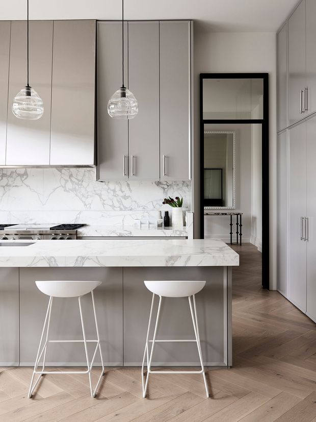 A refined contemporary kitchen with a central island clad in veined white stone, pale grey flat-panel cabinetry, and two white bar stools. Matching stone forms the splashback and benchtop, while globe pendant lights hang above the island. A black-framed doorway leads to a hallway with timber flooring and additional built-in storage.