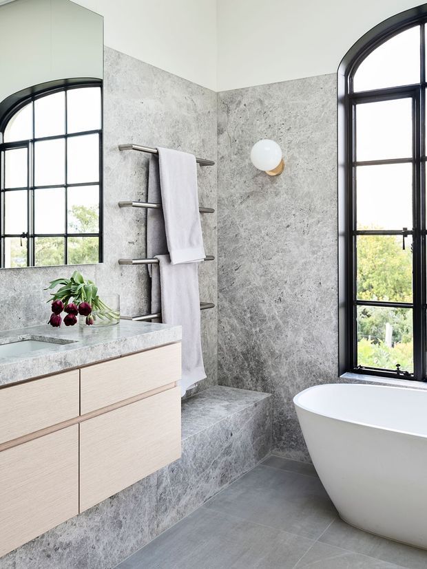 Bright contemporary bathroom with grey veined stone wall and floor finishes, a floating light timber vanity with integrated basin, brushed metal towel rail, freestanding white bathtub, round wall sconce, and tall black-framed arched windows. A mirror reflects the window and vanity area.