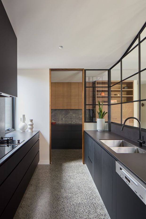 A narrow contemporary galley kitchen with dark matte cabinetry on both sides, integrated appliances, a deep double-bowl stainless-steel sink, and a dark stone-look benchtop. The room has a light terrazzo-style floor, white walls and ceiling, timber-framed cabinetry and a timber-lined pantry or storage zone at the rear. Black-framed glazed partitioning provides visual separation while maintaining openness. Minimal decorative objects and a potted plant add restrained styling.