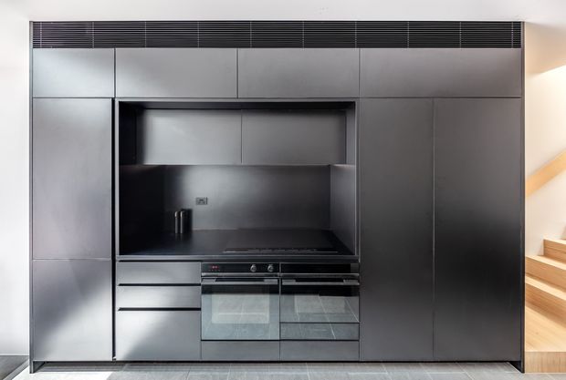 A sleek, fully integrated dark kitchen wall with floor-to-ceiling matte cabinetry, concealed appliances, a black cooktop and oven, and a recessed cooking niche with dark splashback. A timber staircase is visible at the right edge, and a linear ventilation grille runs along the top.
