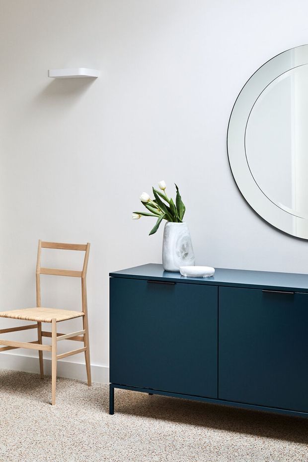 A minimalist interior vignette featuring a dark blue built-in or freestanding sideboard with slim black legs and handles, a light timber folding chair, a white sculptural vase with flowers, and a large round wall mirror. The room has pale walls, a small white wall shelf, and a speckled neutral carpet.