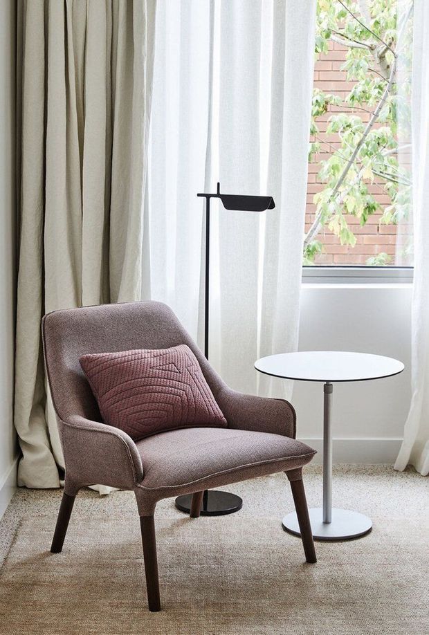 A softly lit reading nook with a mid-century-inspired upholstered armchair, textured cushion, slim black floor lamp, and small round side table. Full-height sheer and beige curtains frame a window overlooking a brick wall and leafy tree. The space has a calm, refined neutral palette and carpeted floor.