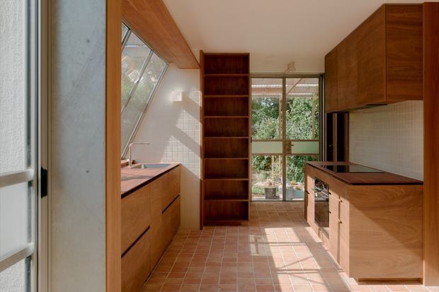 A warm, elongated contemporary kitchen with extensive dark timber cabinetry, a central open shelving unit, terracotta-toned tiled flooring, and a tiled backsplash. Timber-framed glazing and a sloping glazed section bring in abundant natural light, while the kitchen island and parallel work runs create a galley-style layout.