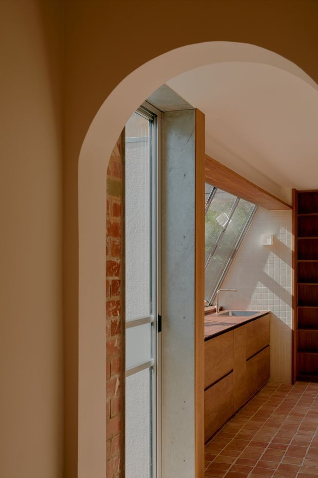 A warm, richly detailed interior view through a rounded plaster archway into a compact kitchen. The kitchen features terracotta tiled flooring, timber cabinetry and shelving, a tiled splashback, exposed brickwork, a concrete-framed window or door opening, and a sloping glazed roof or clerestory window that brings in natural light. The composition highlights the transition between spaces and the mix of earthy materials.