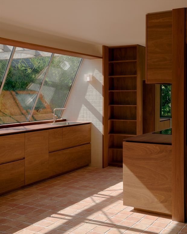 A warm, compact kitchen with extensive walnut-toned timber cabinetry, open shelving, dark benchtops, and a stainless-steel sink. Slanted glazed windows bring in abundant natural light and overlook greenery, while terracotta-toned square floor tiles and white wall tiles create a tactile, earthy interior. The cabinetry and shelving provide strong built-in storage, with a peninsula or island in the foreground.