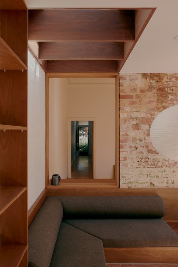 Interior view of a warm, compact living room with a dark upholstered sectional sofa in the foreground, extensive built-in timber shelving along the left wall and timber-lined overhead storage or ceiling elements. A timber-framed internal opening leads to a hallway, while an exposed painted brick feature wall occupies the right side. The space combines natural wood, muted upholstery, pale plaster and aged brick in a refined, inviting scheme.