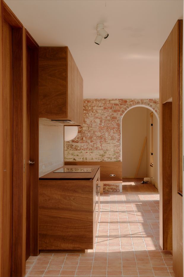 A warm, narrow kitchen corridor with rich timber cabinetry, a compact island or peninsula, pale tiled splashback, terracotta floor tiles, and an exposed red-brick feature wall. An arched doorway leads to an adjacent space, while a white track spotlight provides focused illumination. The design combines original masonry character with clean contemporary joinery.