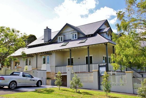 A large traditional multi-storey suburban house with a steeply pitched gabled roof, broad covered front verandah, pale rendered walls, white railings, dormer-style upper windows, and a prominent chimney. The property is photographed from the street with a driveway, front lawn, mature trees, and a parked vehicle in the foreground.