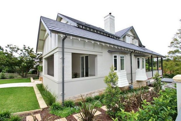 Exterior view of a renovated white weatherboard-style house with a steep pitched metal roof, multiple gabled dormer elements, chimney, covered veranda, and projecting bay windows. The home is set within a landscaped garden with lawn, planting beds, and paved paths.