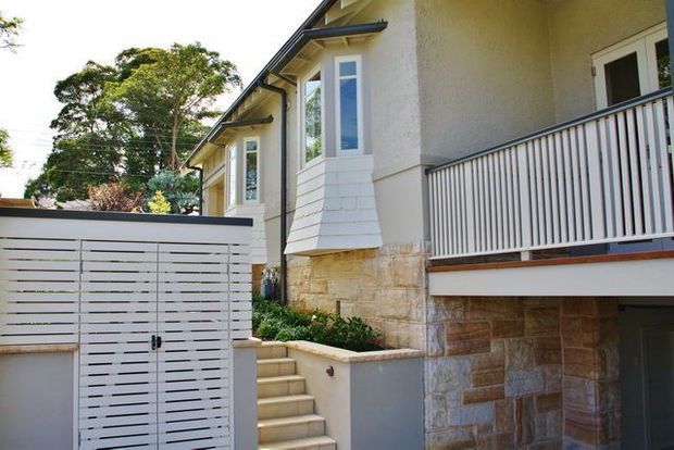 Exterior view of a multi-level residential home with a light rendered facade, projecting bay window, white slatted garage or gate door, balcony railing, stone-clad lower level, and a stairway leading to the entrance. Mature landscaping is visible around the property.
