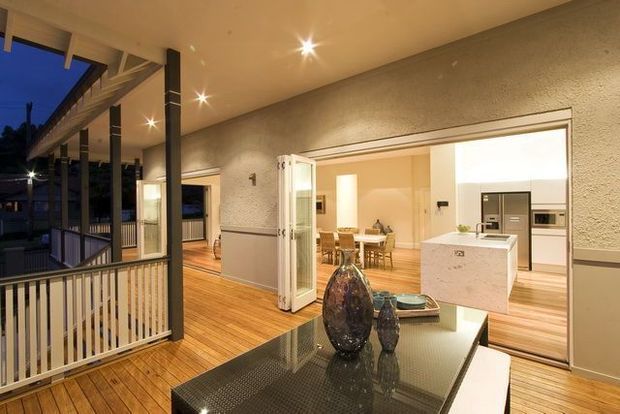 A warmly lit contemporary open-plan interior connecting a covered outdoor terrace or balcony with a dining area and kitchen. Timber decking continues toward the interior, while dark metal posts and railings frame the outdoor edge. Inside, a glossy black dining table with decorative glass vessels sits in the foreground, with a white kitchen island, timber flooring, pale textured walls, and expansive glazed openings visible beyond.