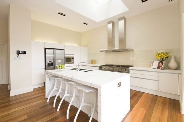 Bright, spacious contemporary kitchen with a large white island and four white bar stools, extensive white cabinetry, stainless-steel appliances, integrated cooktop and rangehood, and warm timber flooring. A skylight and recessed ceiling lights provide abundant illumination, while a vase and small decorative arrangement add subtle styling.