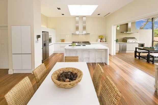 Bright open-plan kitchen and dining area with white cabinetry, a central island with seating, stainless-steel appliances, warm timber flooring, and a white dining table surrounded by woven wicker chairs. Large openings connect the kitchen to a secondary living or outdoor-facing area with abundant natural light.