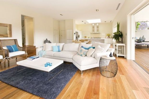 Bright, open-plan living room with a large white sectional sofa, white coffee table, navy area rug, timber flooring, and coastal-inspired blue accents. The room connects visually to a kitchen and dining area, with a wood-lined adjacent space visible through the wide opening on the right. Recessed lighting, skylights, white cabinetry, and indoor plants create an airy contemporary interior.