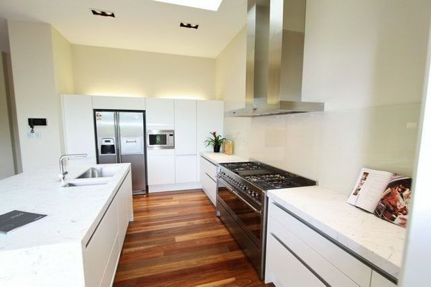 Bright contemporary kitchen with white flat-panel cabinetry, white stone benchtops, stainless-steel appliances and rangehood, a large island with double sink, and warm timber flooring. The space has recessed ceiling lighting and a clean, streamlined layout.