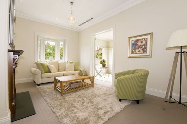 A bright, traditionally detailed living room with pale neutral walls, white decorative trims, a cream sofa, lime-green upholstered armchair, timber coffee table, textured area rug, framed artwork, floor lamp, and a decorative fireplace or mantel at the left. A glazed doorway opens to a small adjoining sunlit space.