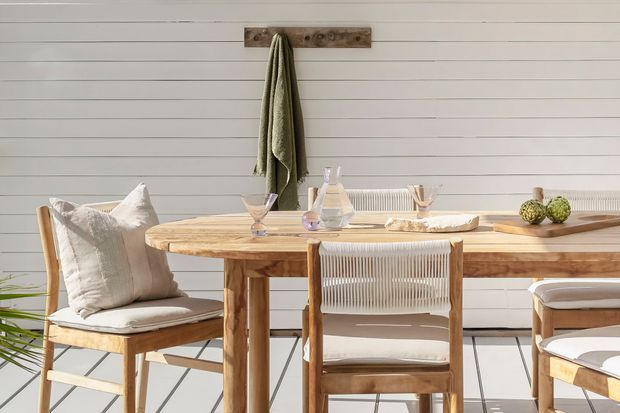 Bright dining area with a rustic timber dining table and chairs against a white horizontal-panelled wall. A wall-mounted timber rack holds a draped textile, while the table is simply set with glassware, a ceramic vessel, a candle, and a small bowl of green fruit. Strong sunlight creates a warm, relaxed atmosphere.