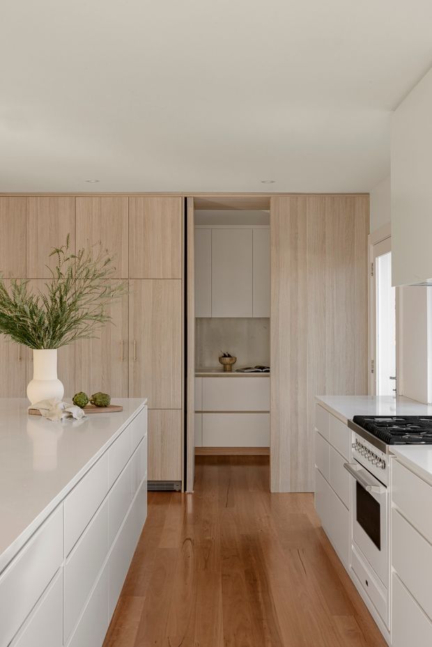 A bright, refined contemporary kitchen viewed down the central aisle. The space features full-height light timber cabinetry, white handleless island cabinetry with a pale solid benchtop, a matching white cooking run with a freestanding-style range, integrated pantry storage, warm timber flooring, and a neutral stone-toned splashback. A vase and small decorative produce sit on the island, while daylight enters from the right.