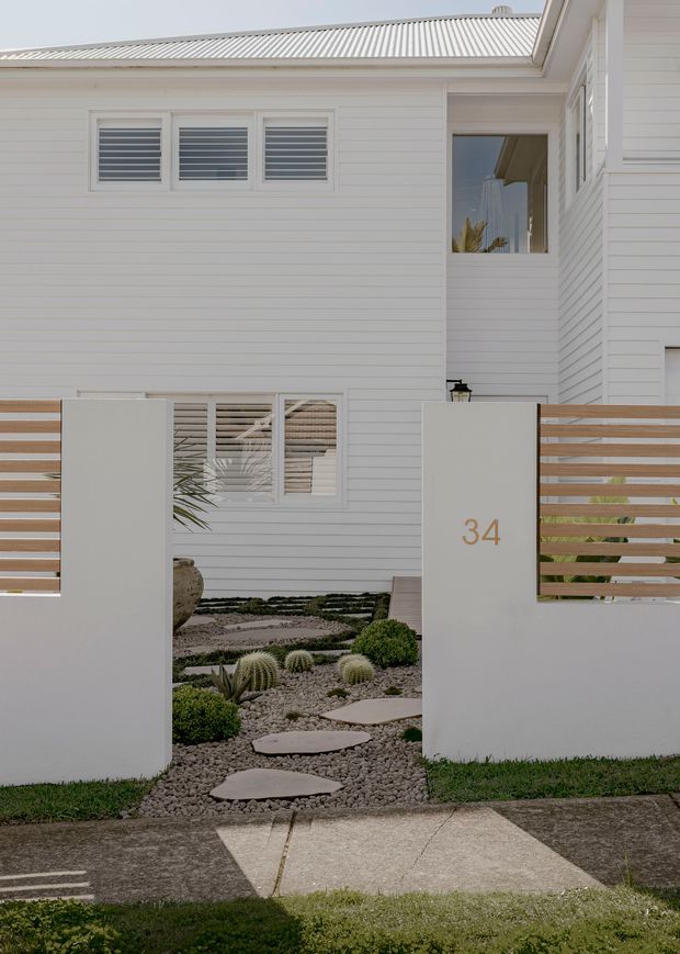 Street-facing view of a contemporary coastal-style residence with white horizontal weatherboard cladding, a light corrugated metal roof, louvred windows, and a low white rendered boundary wall with timber slat screening. A landscaped front entry uses gravel, stepping stones, sculptural succulents, and clipped shrubs, with the house number 34 displayed on the gate pillar.