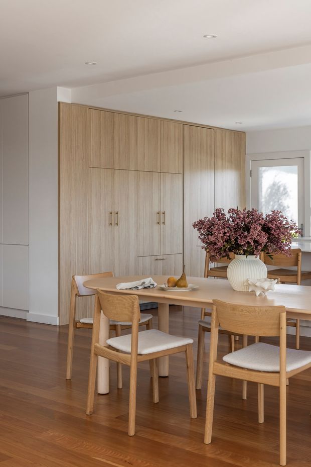 A bright, refined dining area with a large light timber dining table and upholstered timber dining chairs. The room features warm timber flooring, a full-height light timber built-in cabinetry wall with integrated doors and brass-toned handles, white walls and ceiling with recessed downlights, and a windowed rear wall. A sculptural white vase with pink floral branches forms the central table arrangement.