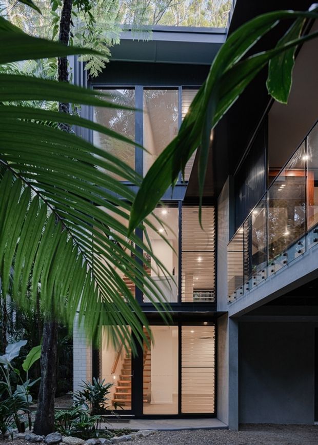 A modern multi-storey house viewed from a leafy landscaped exterior, with dark metal cladding, extensive glazing, an upper-level balcony with glass balustrade, and warm timber-lined interiors visible through the windows. Tropical foliage frames the facade, while a recessed garage or covered area occupies the lower right.