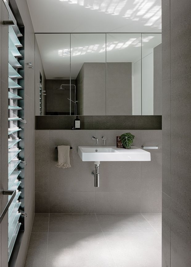 Contemporary minimalist bathroom with a wall-mounted white basin, chrome tapware and exposed plumbing, large grey stone-look wall and floor tiles, mirrored cabinetry, a dark stone horizontal shelf, and louvred glazing allowing filtered daylight. A shower is visible in the mirror reflection.