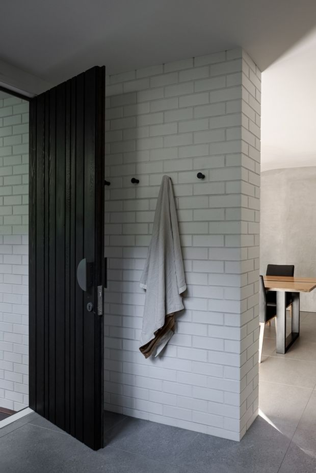 A contemporary bathroom or shower entry with white painted brick wall tiles, dark grey floor tiles, matte black vertical slatted door or screen, and black wall hooks holding a towel. A dining area with a timber table is visible beyond the opening.