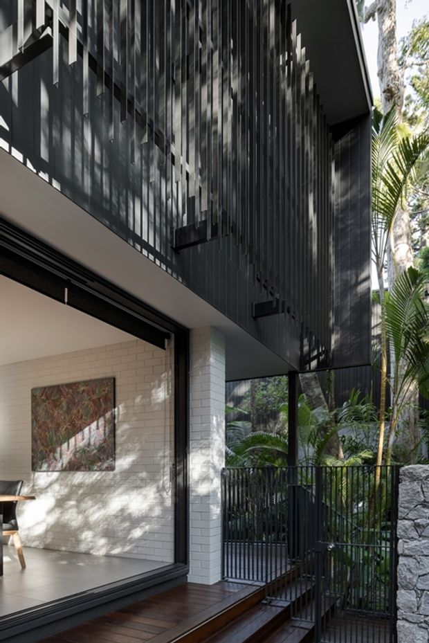 A close architectural view of a contemporary multi-level house in a lush tropical setting. The facade combines dark vertical metal screening and fins with white masonry, expansive black-framed glazing, a timber deck and black metal railings. A partially visible interior with pale brick walls opens onto the deck, while dense palms and stonework frame the entry.