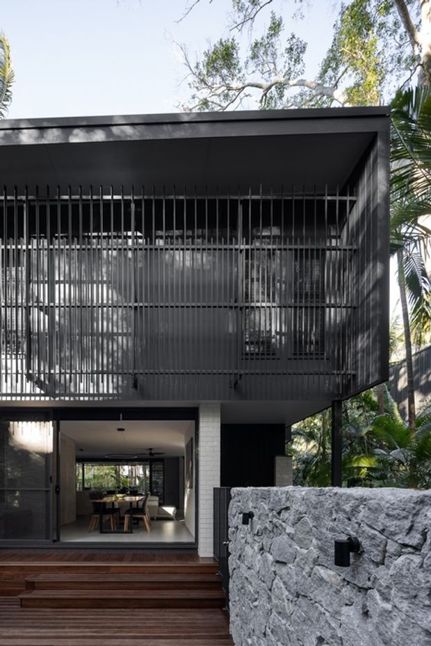 A contemporary two-storey house facade viewed from the front garden, featuring a dark flat-roofed upper level screened with closely spaced vertical metal battens. The lower level has large glazed openings leading to a dining area, dark timber decking and steps, and a rough pale stone retaining wall with integrated black wall lights. Dense tropical landscaping surrounds the building.