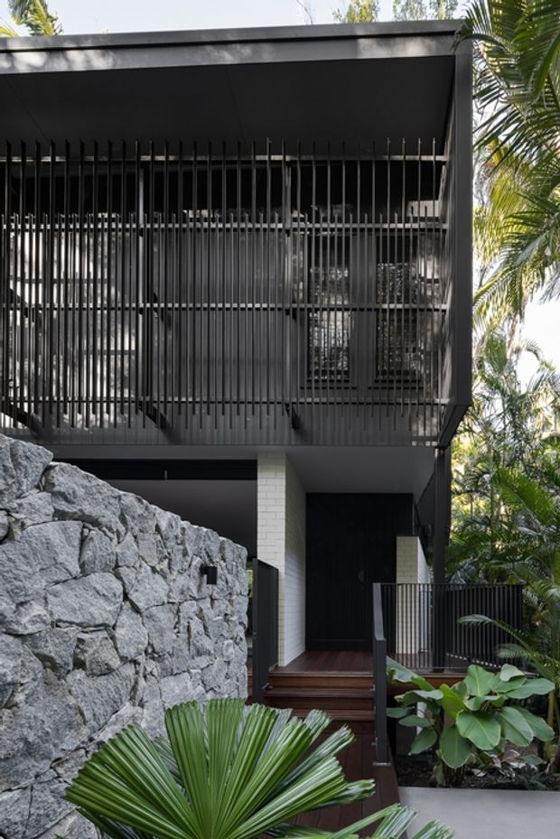 A modern multi-level residential facade in a lush tropical setting. The dark rendered or metal-clad structure features a projecting upper balcony screened by closely spaced vertical black battens, dark-framed glazing, a flat roofline, timber steps and decking, and a white masonry column. A rugged natural-stone retaining wall and tropical planting frame the entry.
