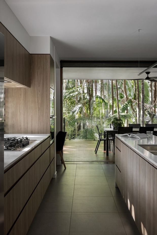 A contemporary galley kitchen with two parallel banks of dark timber-grain cabinetry and pale stone benchtops. A gas cooktop is integrated into the left run, while the right run includes a sink and raised breakfast or dining counter. The kitchen opens directly onto a timber deck and lush tropical garden through wide glazing, with a dining table, ceiling fan and pendant light visible beyond. Large-format grey floor tiles and a restrained neutral palette create a refined, indoor-outdoor atmosphere.