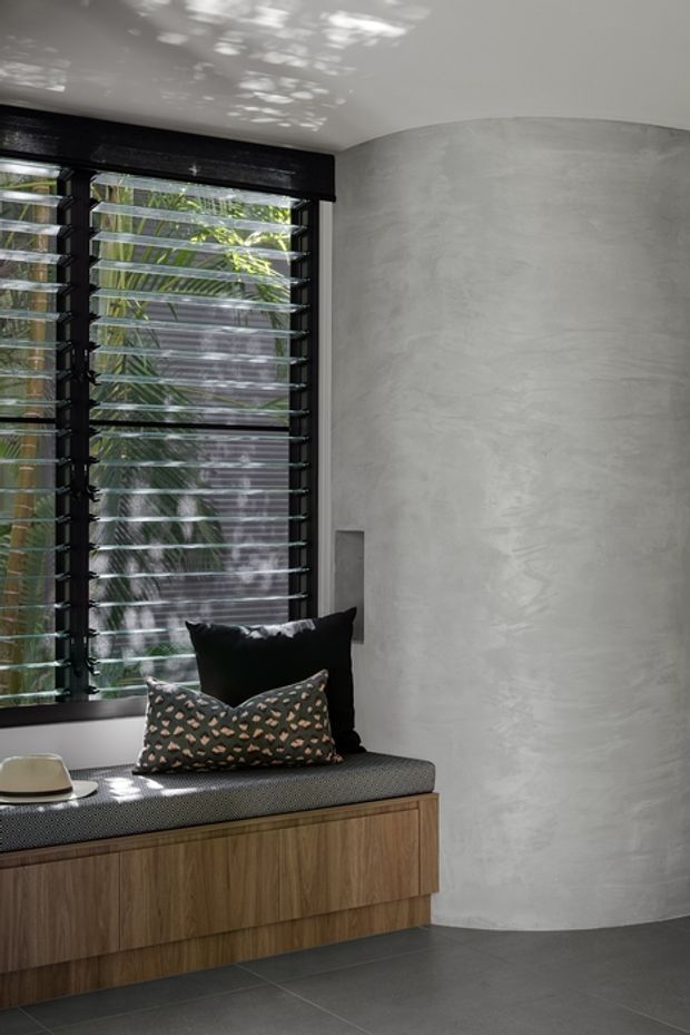 A refined built-in window seat with dark upholstered cushioning and layered cushions sits above warm timber cabinetry. The space features a large black-framed louvred window, smooth light-grey curved plaster walls, and large dark-grey floor tiles. Dappled daylight and tropical greenery are visible through the glazing, creating a calm, contemporary interior nook.