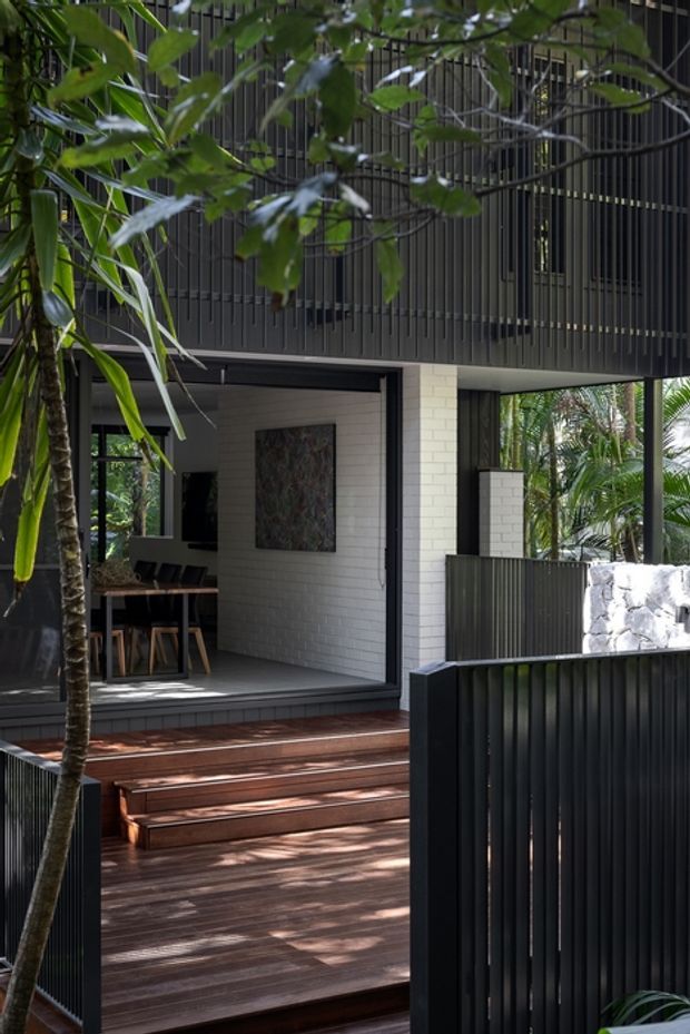 A contemporary residential exterior viewed from a landscaped outdoor approach, featuring dark vertical slatted cladding and fencing, a raised timber deck with broad steps, black-framed glazing, and a pale brick wall. A dining area is visible through the open glazing, surrounded by lush tropical planting and a stone boundary wall.