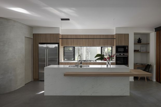 Contemporary open-plan kitchen with a large white stone island and timber breakfast ledge, dark grey flooring, timber cabinetry, integrated stainless-steel refrigerator, built-in oven and microwave, and a concrete-look curved feature wall. Horizontal windows with external louvres bring in filtered daylight, while open shelving and a small dining bench are visible to the right.
