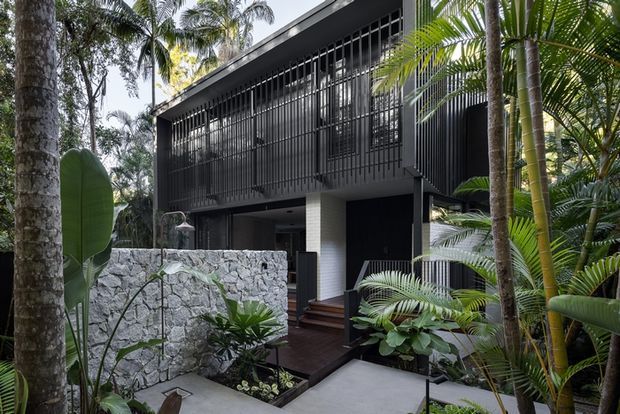 A contemporary multi-level house nestled in lush tropical landscaping. The dark, rectilinear facade features extensive vertical metal screening, dark cladding, and a recessed entry above a pale stone boundary wall. Concrete paths, timber steps, dense palms, and broad-leaf planting create a secluded garden approach.