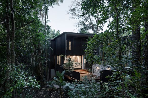 A compact two-storey dark-clad dwelling nestled within dense native bush. The home features a simple flat-roofed rectangular form, large glazed openings, a sheltered upper-level balcony or covered outdoor space, and a warm illuminated lower-level living area connected to a small timber deck. Lush vegetation closely surrounds the building.