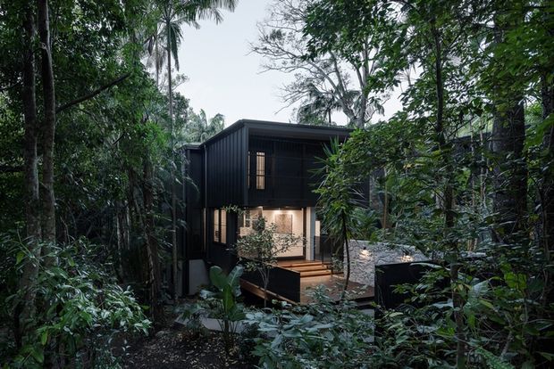 A dark, contemporary multi-level dwelling nestled within dense tropical forest. The building features black vertical cladding, a projecting upper volume, large glazed openings, and timber decking with outdoor seating visible at the lower level.