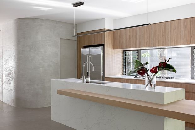Contemporary kitchen interior with a large white stone island and integrated timber breakfast ledge, warm timber overhead cabinetry, a stainless-steel refrigerator, and a textured concrete-look curved wall. Horizontal windows with louvred blinds bring in natural light, while a linear pendant provides task lighting. The palette is warm and neutral with light wood, white, and grey finishes.