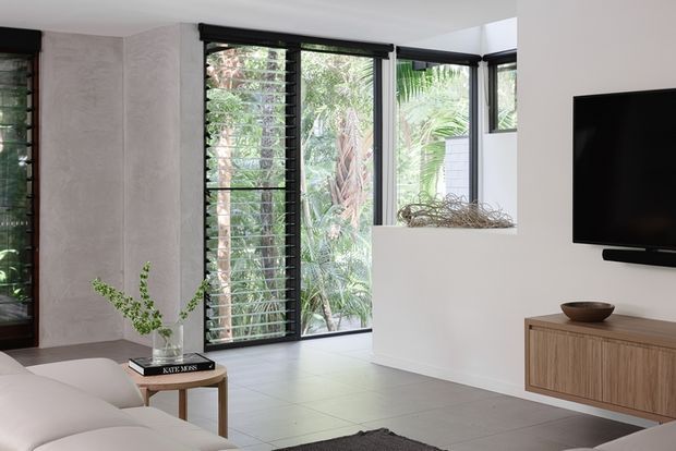 Bright contemporary living room with a white plastered interior, pale tiled floor, large black-framed louvred windows overlooking lush greenery, a wall-mounted television, and a timber media console. A neutral sofa and small side table complete the calm, minimalist setting.