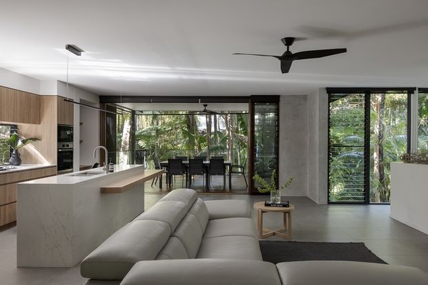 Contemporary open-plan kitchen and living area with a large marble-look island, warm timber cabinetry, light upholstered sectional sofa, dining area, and extensive black-framed glazing overlooking lush vegetation. A low-profile black ceiling fan and recessed lighting add a streamlined finish.