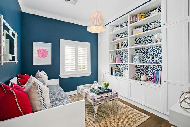 A bright, compact sitting room or reading nook with a white daybed, layered patterned and vivid red cushions, dark teal-blue walls, white plantation shutters, and extensive built-in white shelving and cabinetry. The shelving features bold blue-and-white patterned backing and books, while a woven pendant light and natural-fibre rug add texture. The room has a cheerful, eclectic coastal-inspired character.