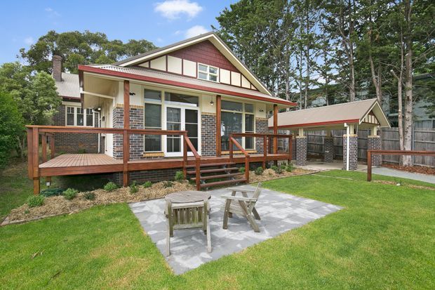 Exterior view of a detached residential house with a prominent front-facing gable, red brick walls, cream weatherboard and trim, and a red metal roof edge. Timber decks, verandas, railings, stairs, and a paved outdoor seating area occupy the landscaped foreground. A separate small gabled outbuilding or carport is visible to the right, with mature trees surrounding the property.