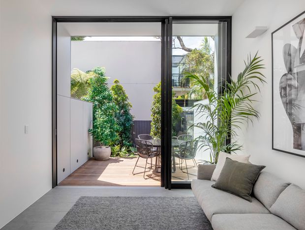 Bright contemporary living room opening through large black-framed glazed sliding doors to a compact timber deck and landscaped courtyard. The room has white walls, a grey rug, a light neutral sectional sofa, indoor palms, and large monochrome wall art. The outdoor area includes café-style seating and privacy walls, with neighbouring buildings visible beyond.