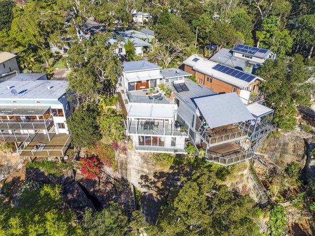 Aerial view of a cluster of contemporary homes built among dense trees on a steep, rocky site. The houses feature light-coloured metal roofs, extensive glazing, balconies and terraces, timber and rendered facades, and several rooftop solar-panel arrays. Elevated decks and stairways connect the homes to the rugged landscape.